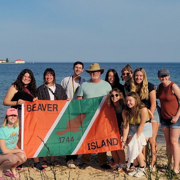 group of 11 people in two rows holding an orange and green flag, Beaver Island 1744 with an image of a beaver in the middle of the flag. They are standing in front of Lake Michigan on shoreline.
