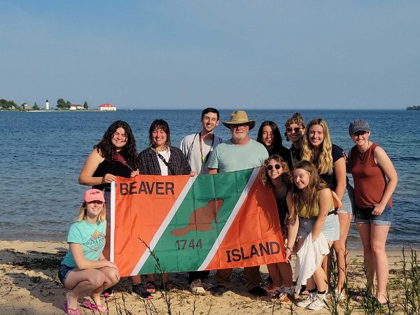 group of 11 people hold Beaver Island flag while standing on shoreline of Lake Michigan
