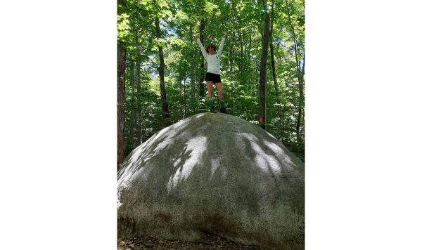 woman stands on top of round, tall rock in forest; she is wearing a white tshirt