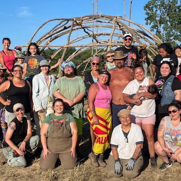 large group of people standing in front a teaching lodge, which is a sacred space to Native Americans constructed from saplings.