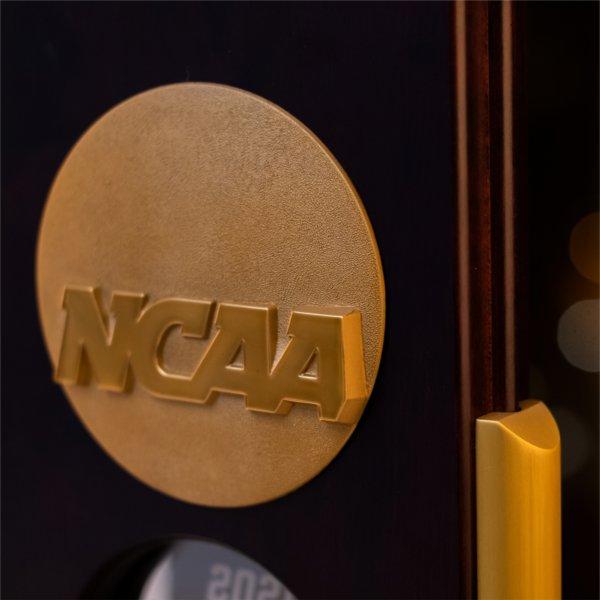 Close-up of an NCAA championship trophy, showing the gold NCAA medallion and part of the wooden frame, with blurred warm lights in the background.