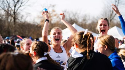 A group of women&#8217;s cross country runners celebrate together outdoors, smiling and cheering with their arms raised after a race, with tents and trees blurred in the background.