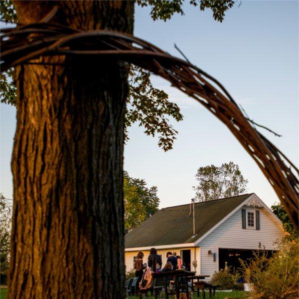 A group of people assemble in front of a white garage. In the foreground, a large wreath of braided branches leans against the trunk of a tree.