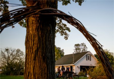 A group of people assemble in front of a white garage. In the foreground, a large wreath of braided branches leans against the trunk of a tree. 
