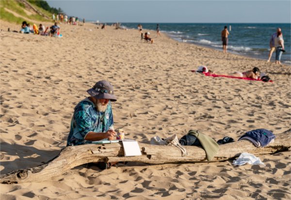 A person wearing a hat sits on a beach next to Lake Michigan, resting a notebook of paper on a log to paint.