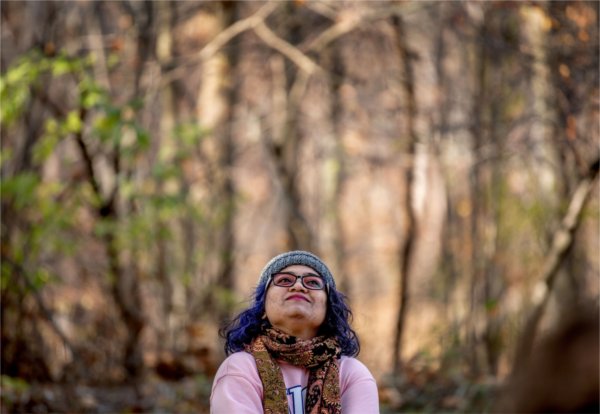 A person wearing a stocking cap and scarf looks upward while sitting amid trees.
