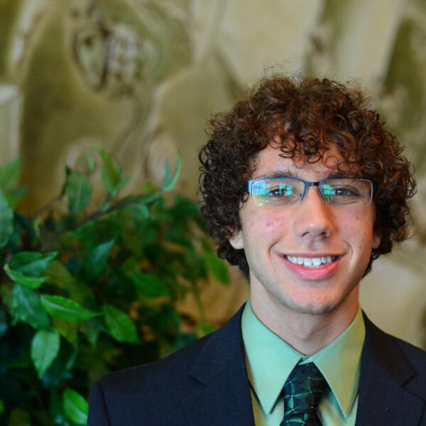 Cameron Jones poses for a photo in a dark blue suit with a light green shirt and dark green tie.