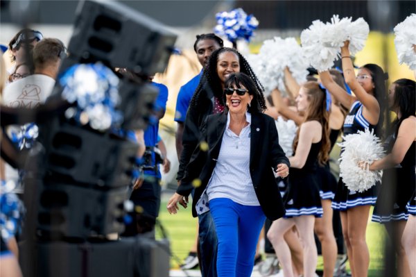 GVSU President Philomena V. Mantella, wearing sunglasses, smiles while cheerleaders wave white pompoms.