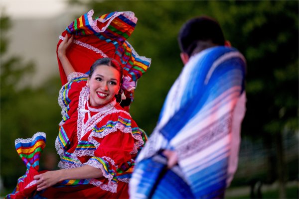 A person wearing a red dress with multi-colored trim smiles at another person while dancing. The person is holding up the train of a skirt while dancing.