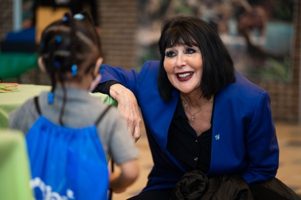 A smiling university president wearing blue kneels to talk to a small child.