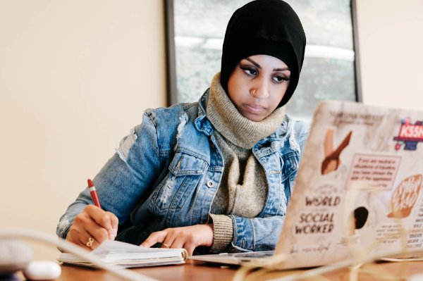 A college student wearing a head covering works on a laptop in a library setting.  