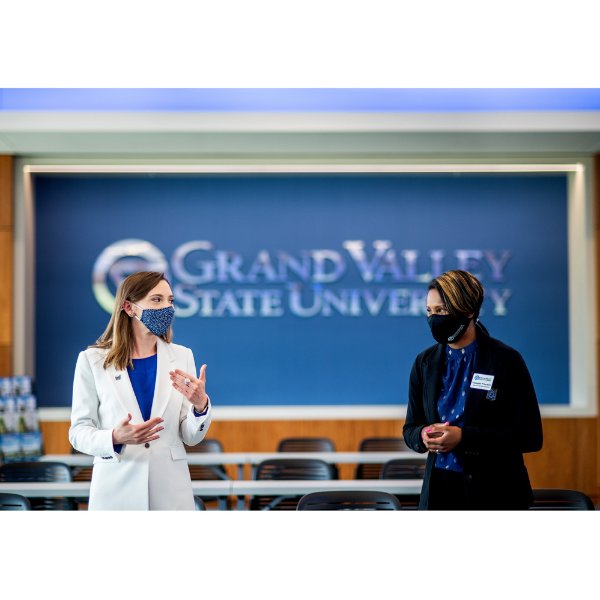 At left, Sarah Tibbe and Maegan Frierson stand and talk to each other in the Laker Experience Suite. Behind them is a blue wall with GVSU logo and words: Grand Valley State University