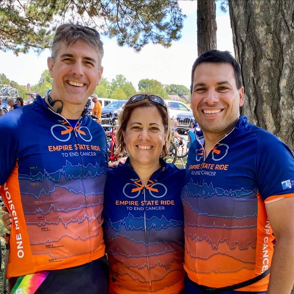trio standing in bicycle shirts that designate Empire State Ride in bright blue and orange, standing in front of a tree