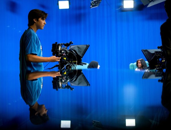A high school student is reflected in a glass table as he runs a television camera. 