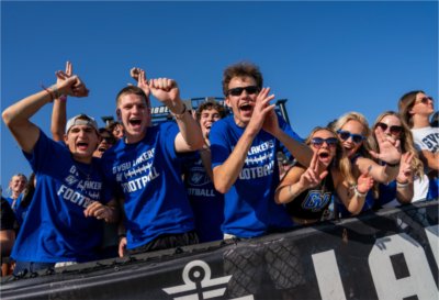 A group of enthusiastic Grand Valley State University fans wearing blue GVSU football shirts cheer and smile from the stands during a sunny game day.
