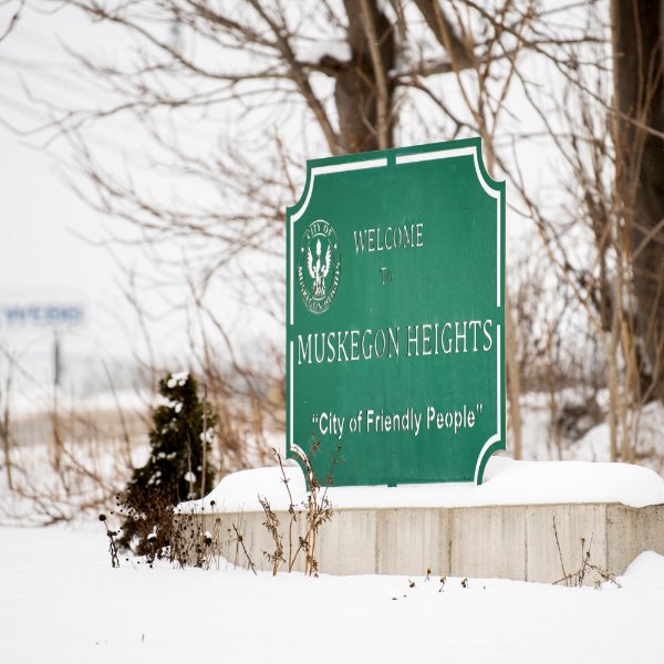A green sign sitting on a raised, snow-covered cement base says: "Welcome. Muskegon Heights. City of Friendly People."