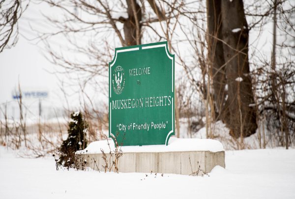 A green sign sitting on a raised, snow-covered cement base says: "Welcome. Muskegon Heights. City of Friendly People."