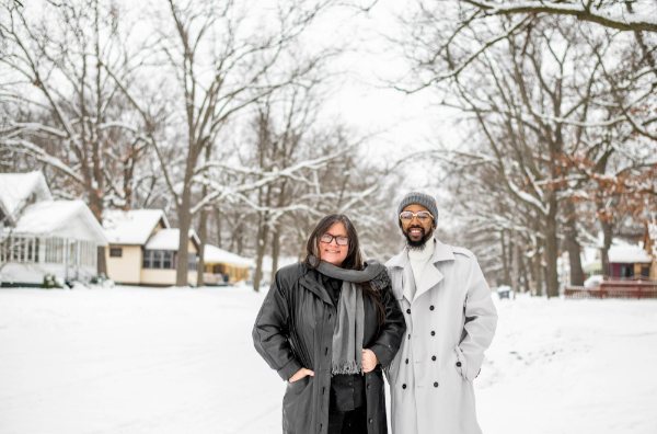 Two people smile while standing on a snow-covered street with houses in the background.