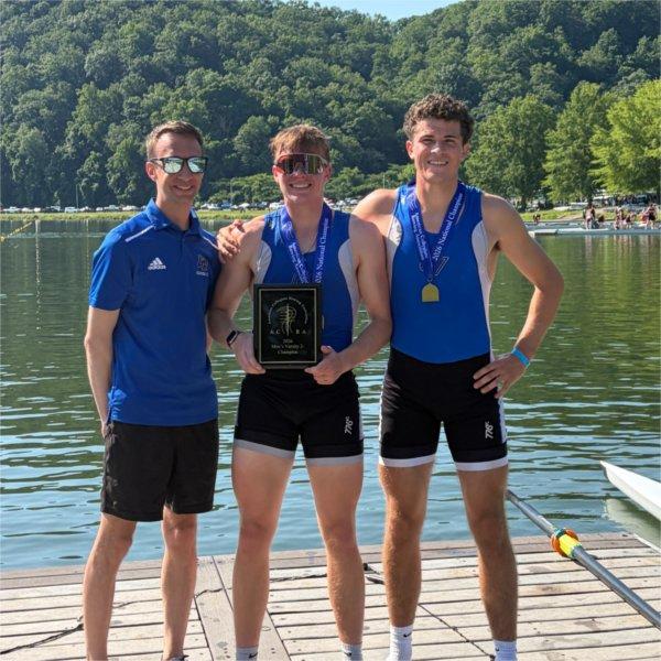 Kyle Barnhart, left, is standing on a dock next to two GVSU rowers, one holding a plaque