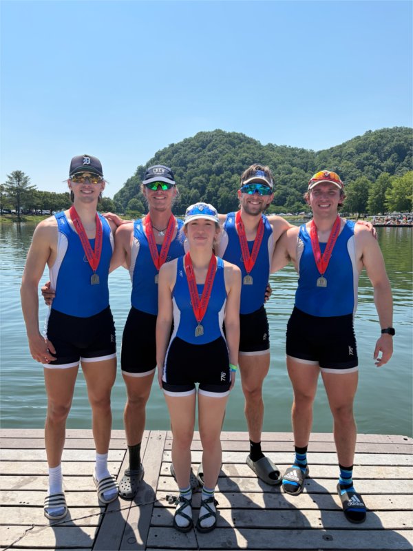 four rowers and a coxswain standing on a dock, with medals around their necks
