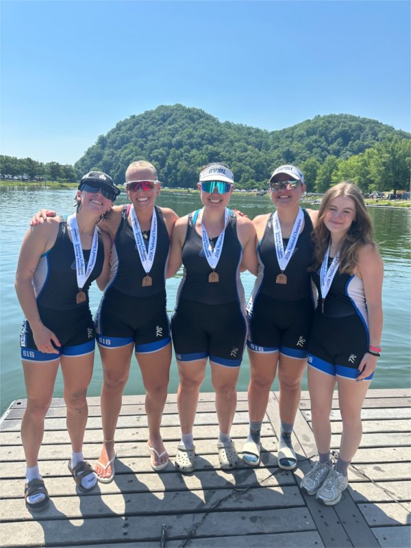 five women rowers standing on a dock with medals, some wear hats and sunglasses