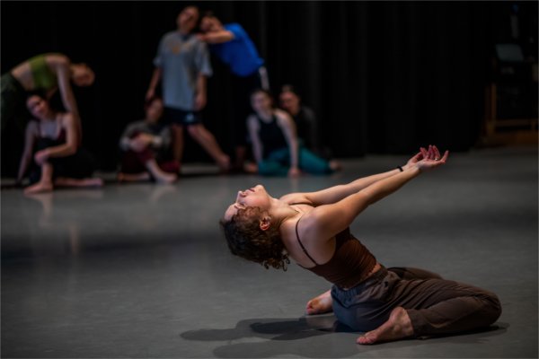 A dancer is on their knees, bending back with arms reaching out. Other dancers are in the background.