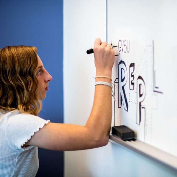 A student draws on a whiteboard with the words GVSU and REP4.