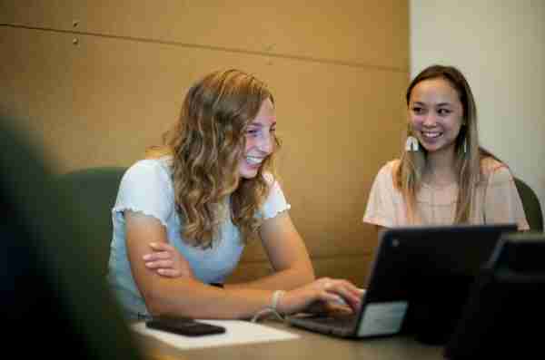 Two students smile one one works on a computer and another sits to the side