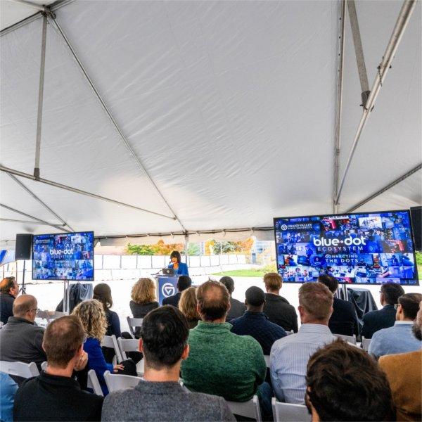 A group of people watch Grand Valley President Philomena V. Mantella speak. Screens with the words, "Blue Dot Ecosystem" and "connecting the dots," along with the GVSU logo, are in the background.