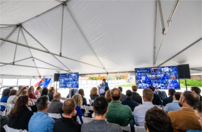 A group of people watch Grand Valley President Philomena V. Mantella speak. Screens with the words, "Blue Dot Ecosystem" and "connecting the dots," along with the GVSU logo, are in the background.