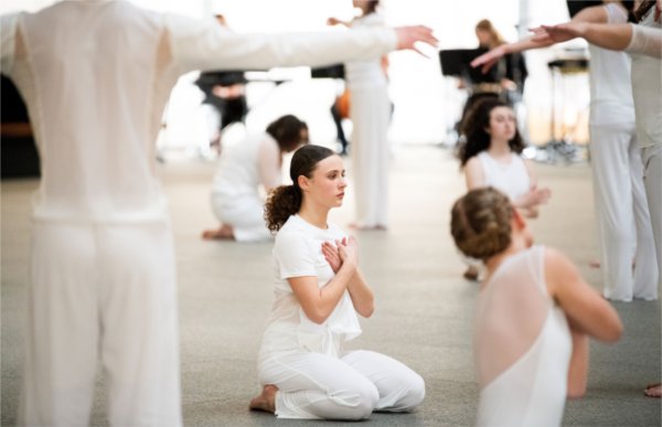  A dancer holds their hands to their heart while surrounded by other dancers wearing white. 