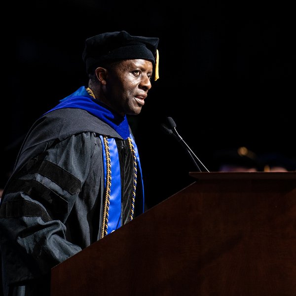 Felix Ngassa in academic regalia speaks at a podium during a commencement ceremony.