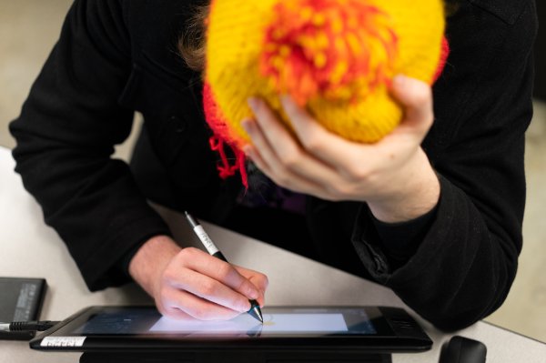 A student works on a piece of art by using a writing instrument and a screen. The student's hand is at the forehead.
