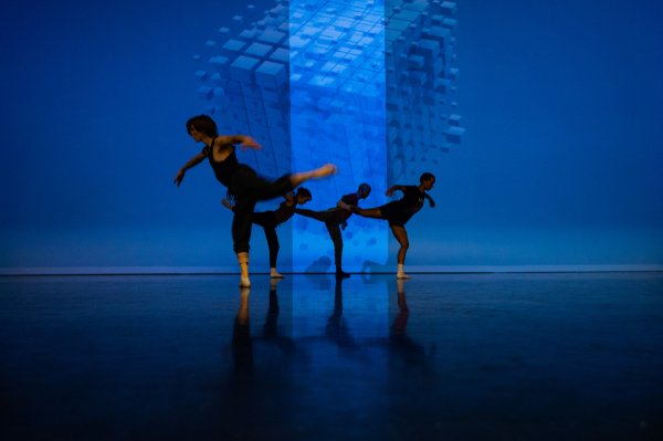 Four dancers perform on stage against a cube projected onto a blue backdrop.