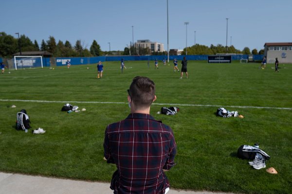 Dr. David Burkard, with back shown, watches the track team practice on the soccer field.