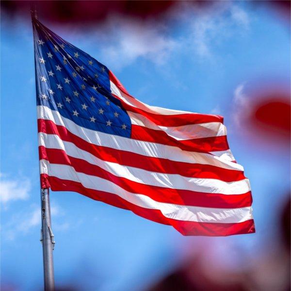 An American flag waves atop a flagpole on Grand Valley's campus against a blue sky with scattered white clouds.