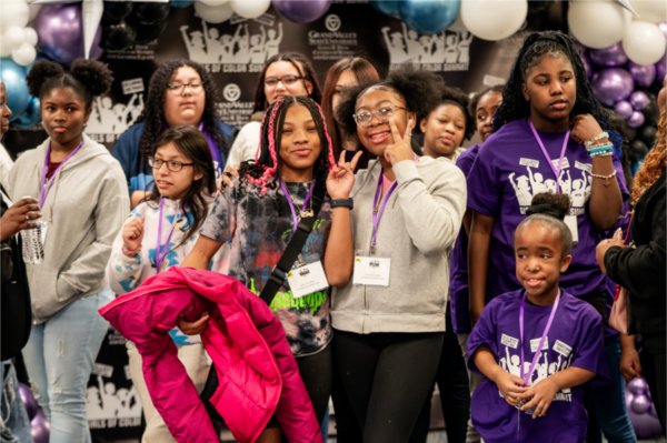 A group of girls pose for a photo in front of a Girls of Color background. The girl in the middle has pink braids and a Goosebumps shirt.