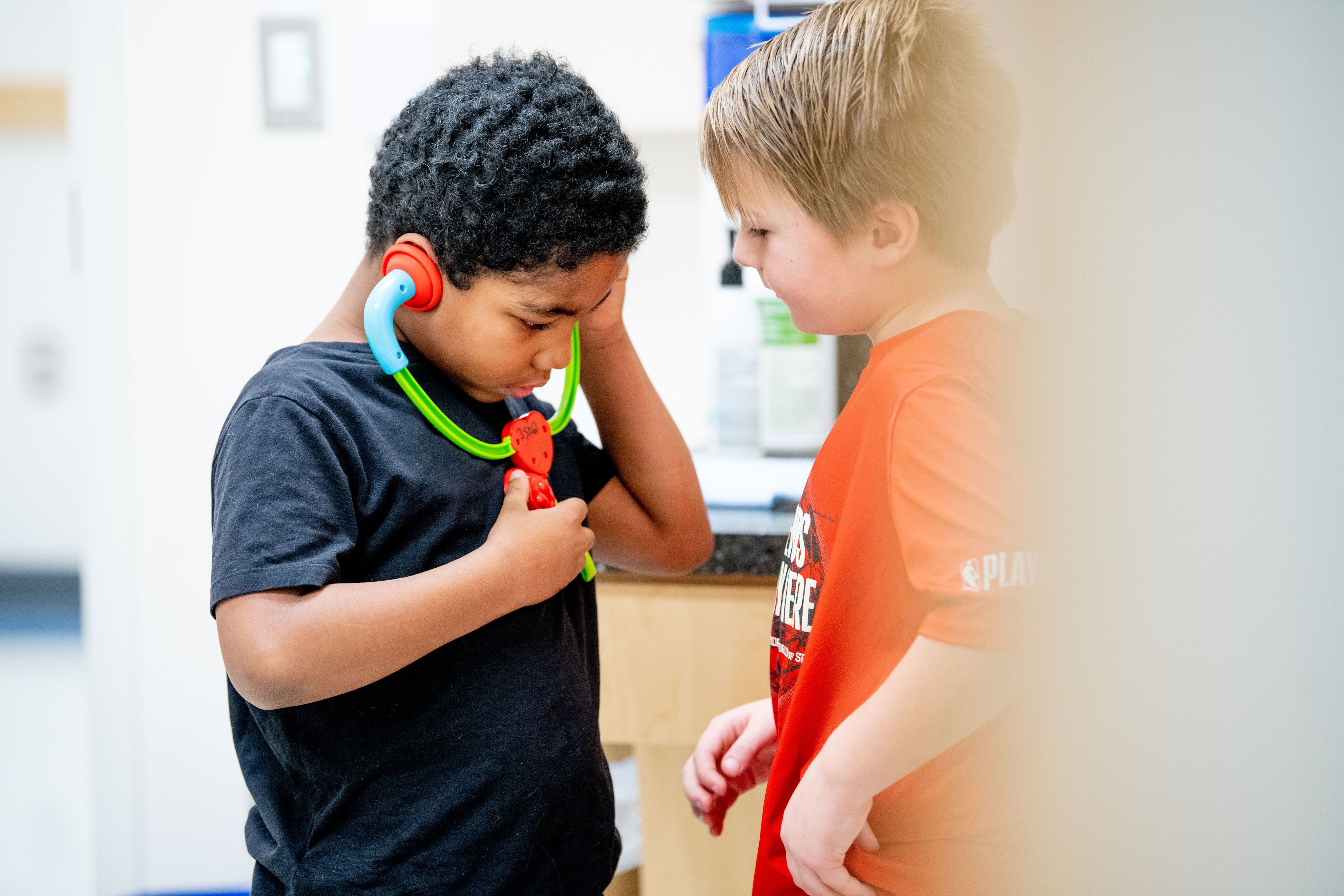 A child plays with a toy stethoscope while another child stands in front watching.
