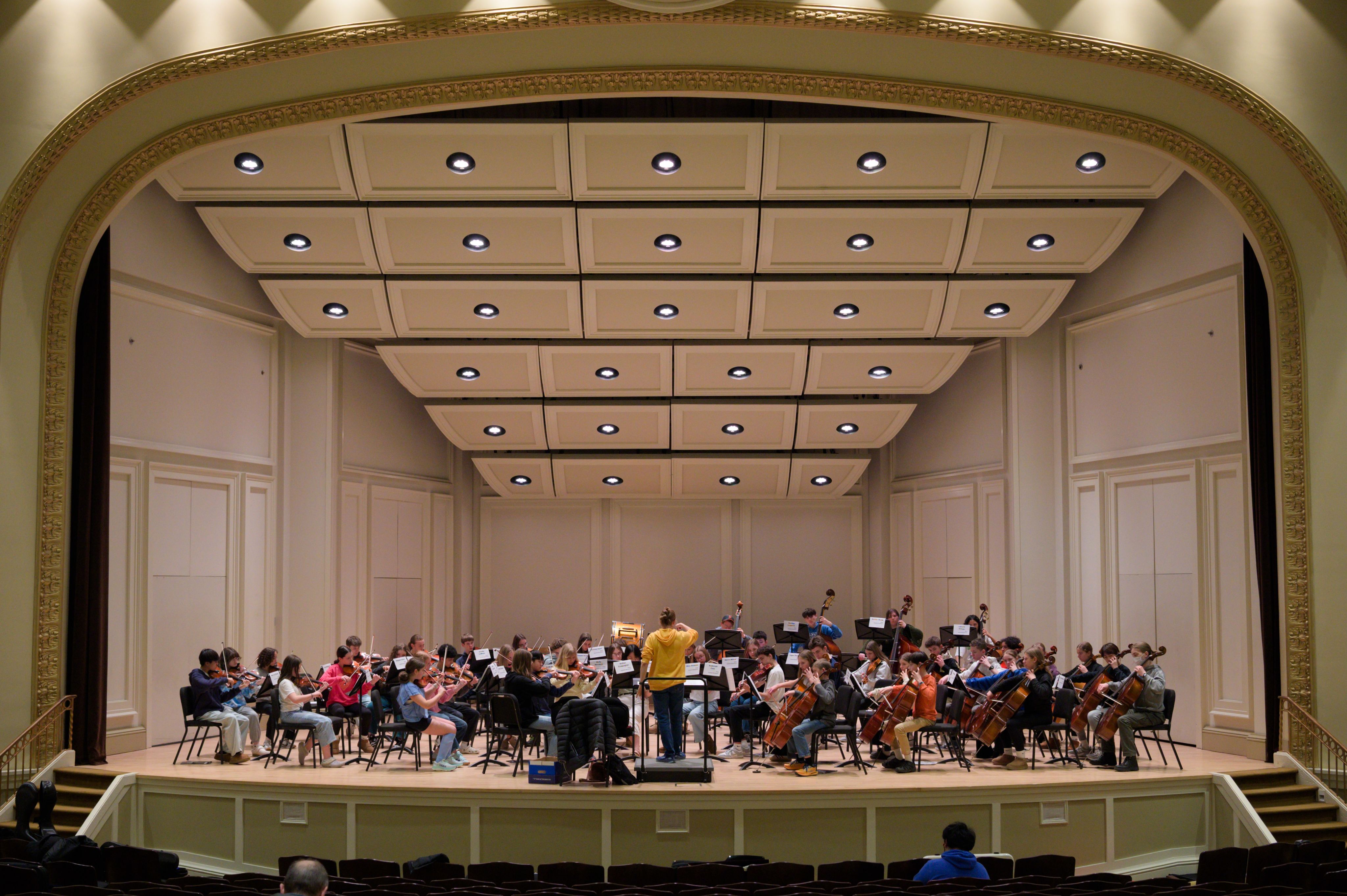 orchestra on stage with condutor in yellow sweatshirt