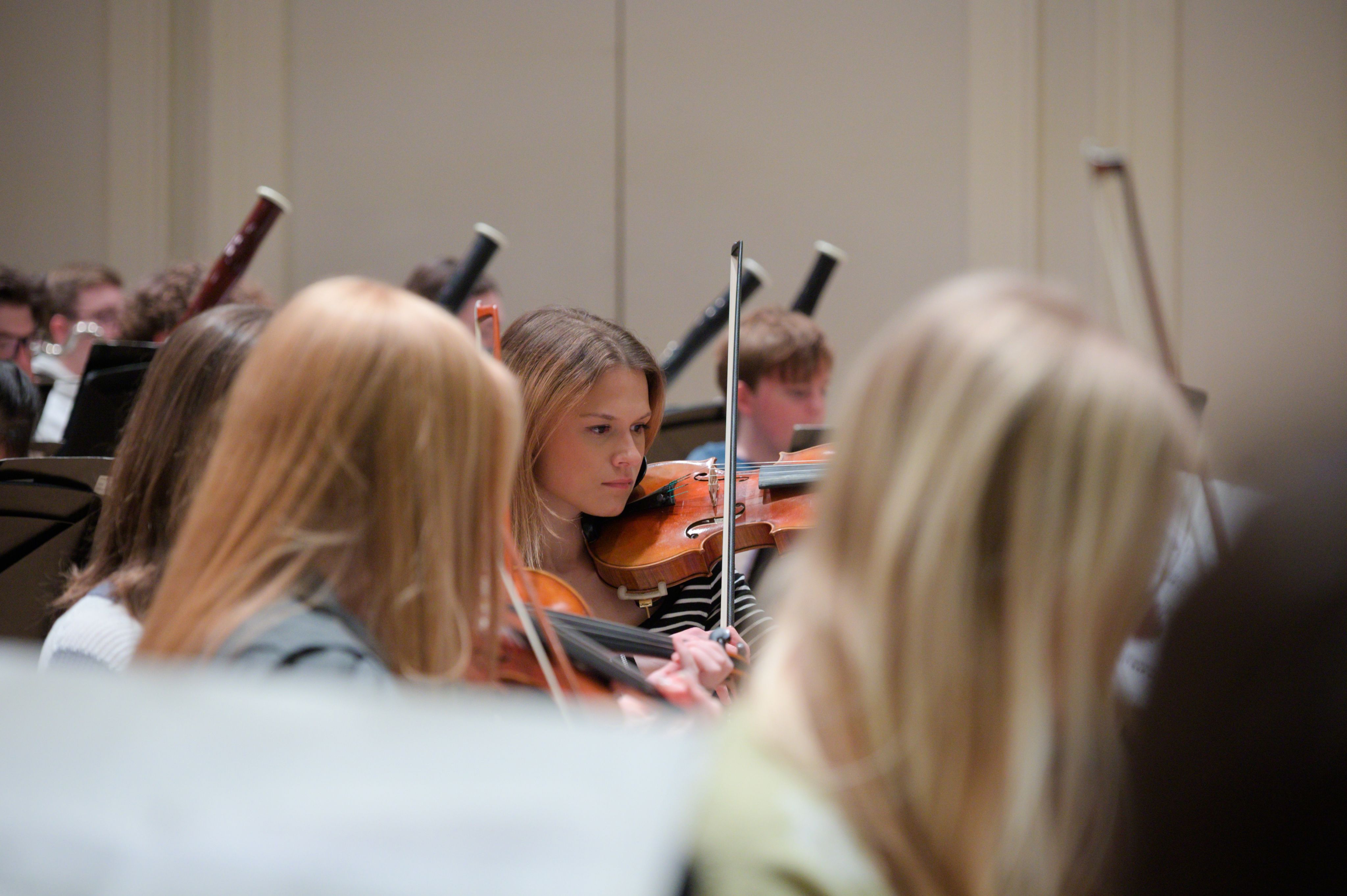 close up of student playing violin, with other instruments behind her in an orchestra on stage