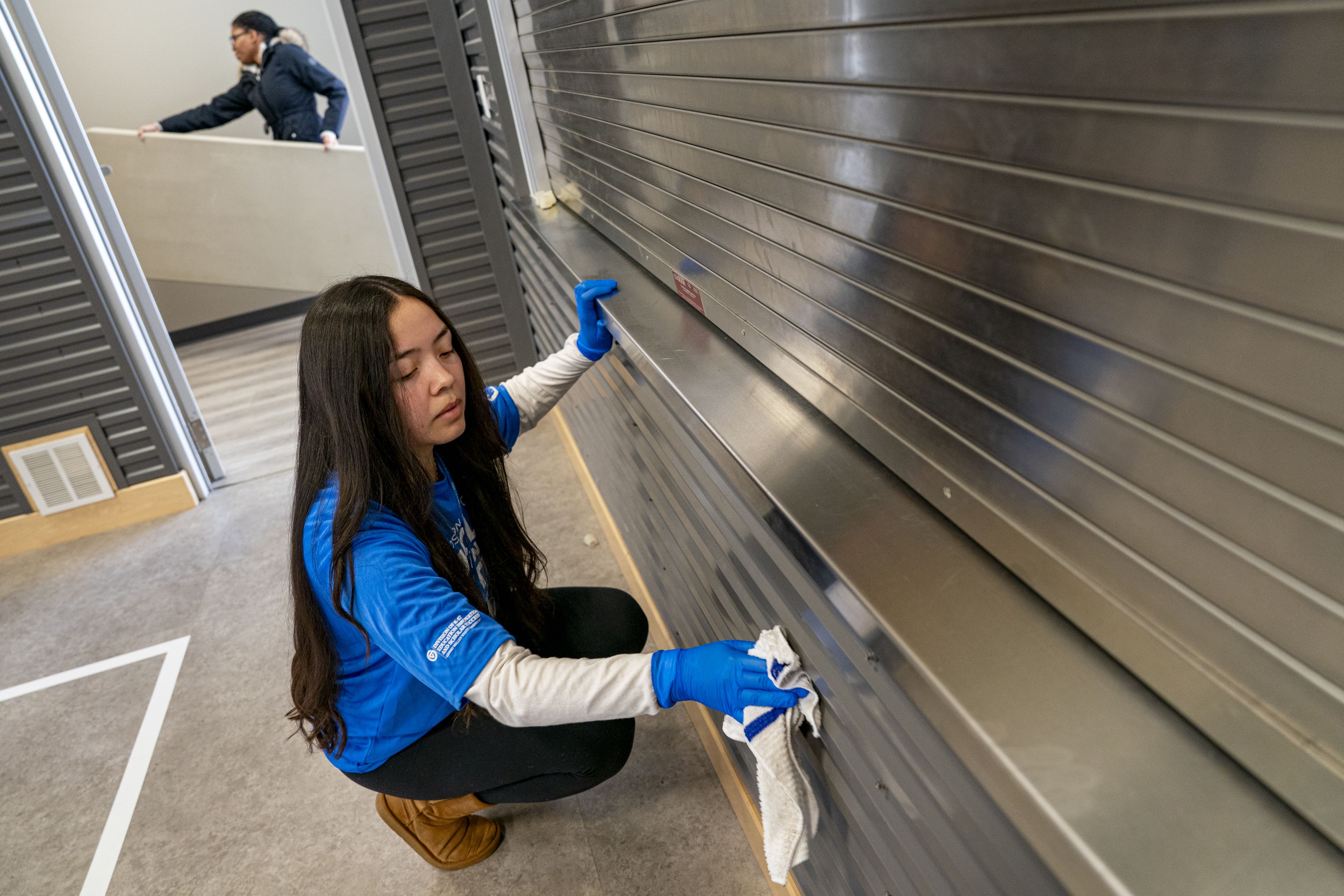 student in blue tshirt kneeling to clean near the floor, another student hauls away a long table