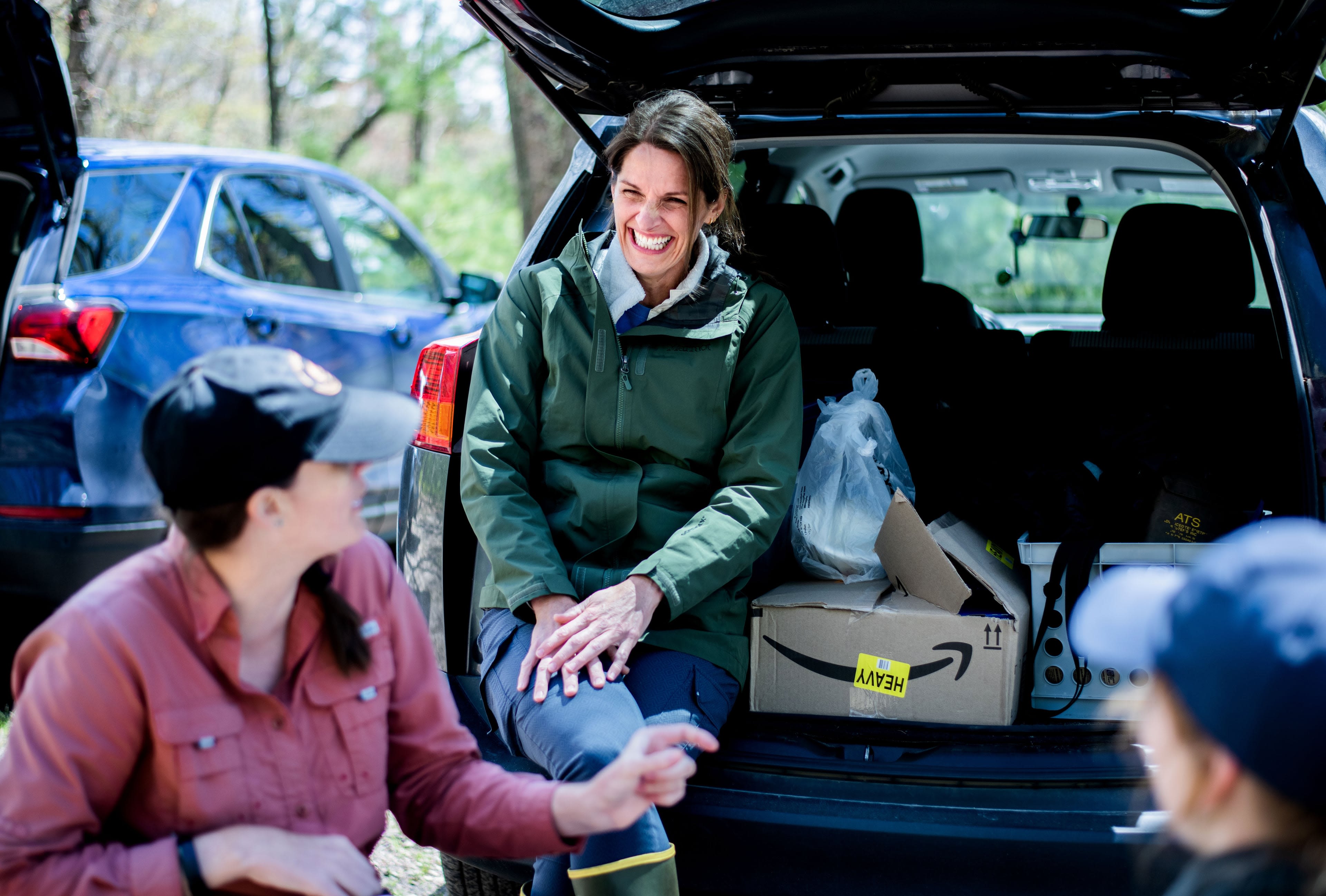 Three people gather around an open SUV trunk filled with boxes. One person inside the trunk smiles, while another gestures nearby.
