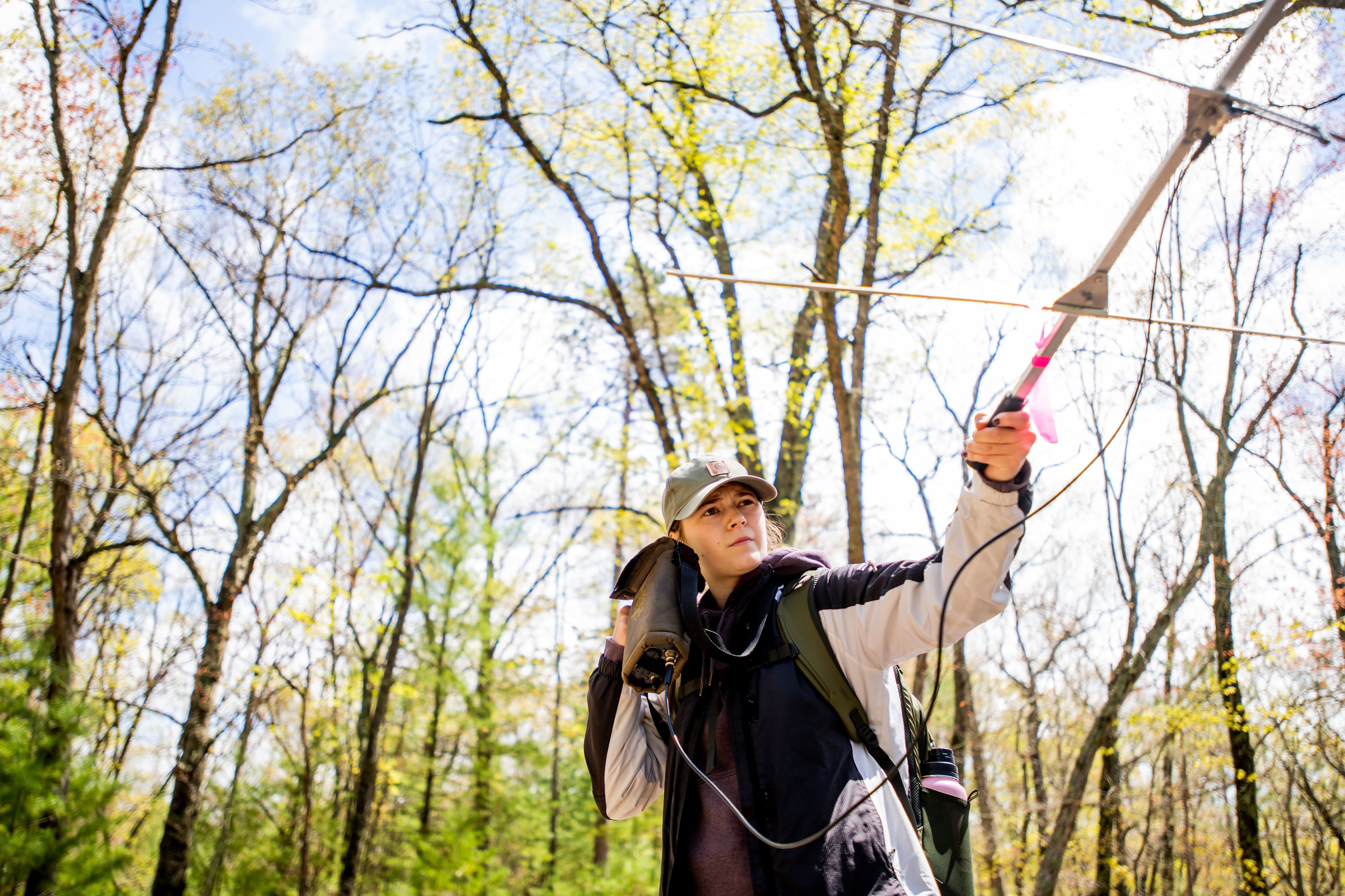 A person in outdoor gear stands in a forest holding a radio telemetry receiver. Tall trees and a bright sky fill the background.