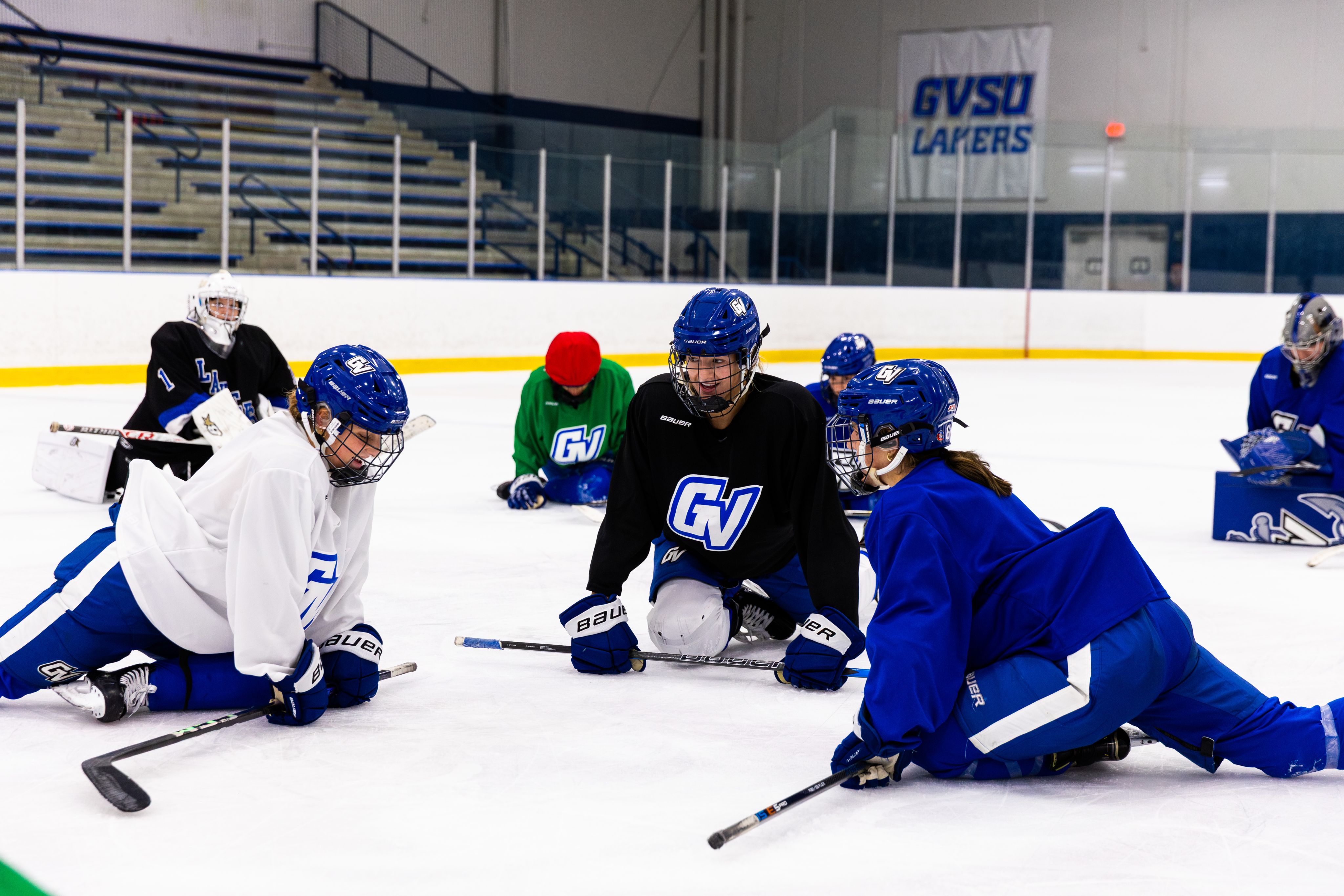 Several people in hockey gear sit on an ice rink stretching their legs.