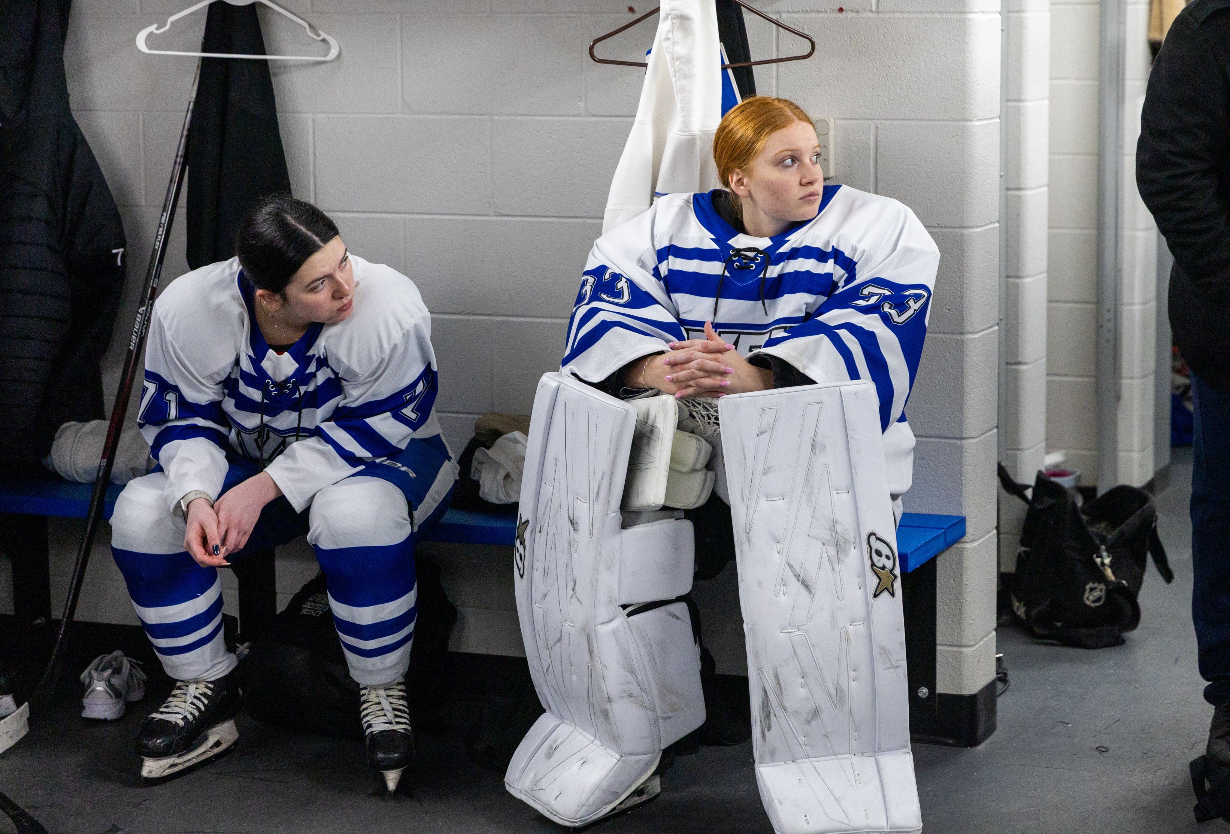 Two hockey players sit on a locker room bench looking off to the right.