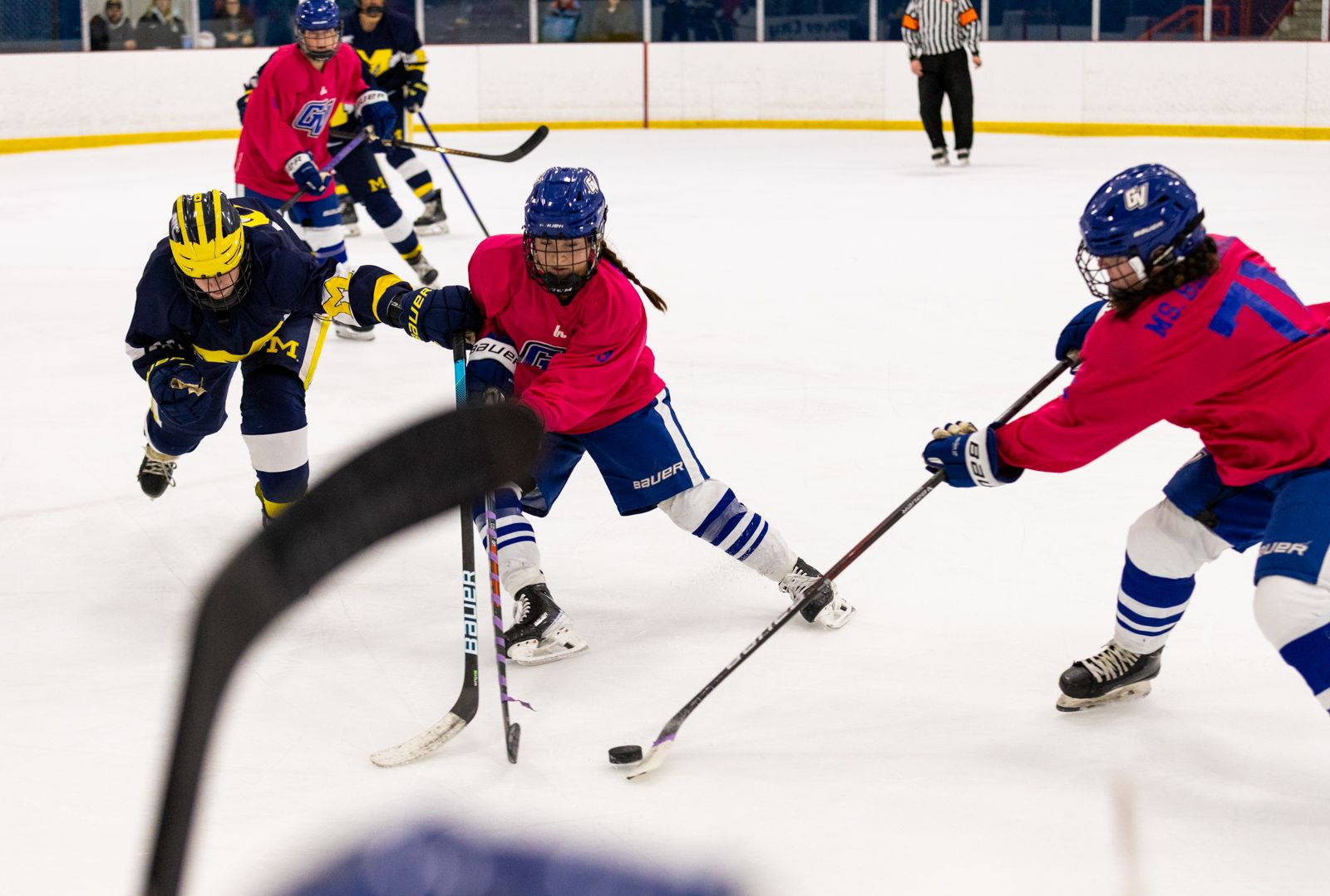 Three hockey player, two in pink jerseys and one in navy, reach for a hockey puck with their sticks.
