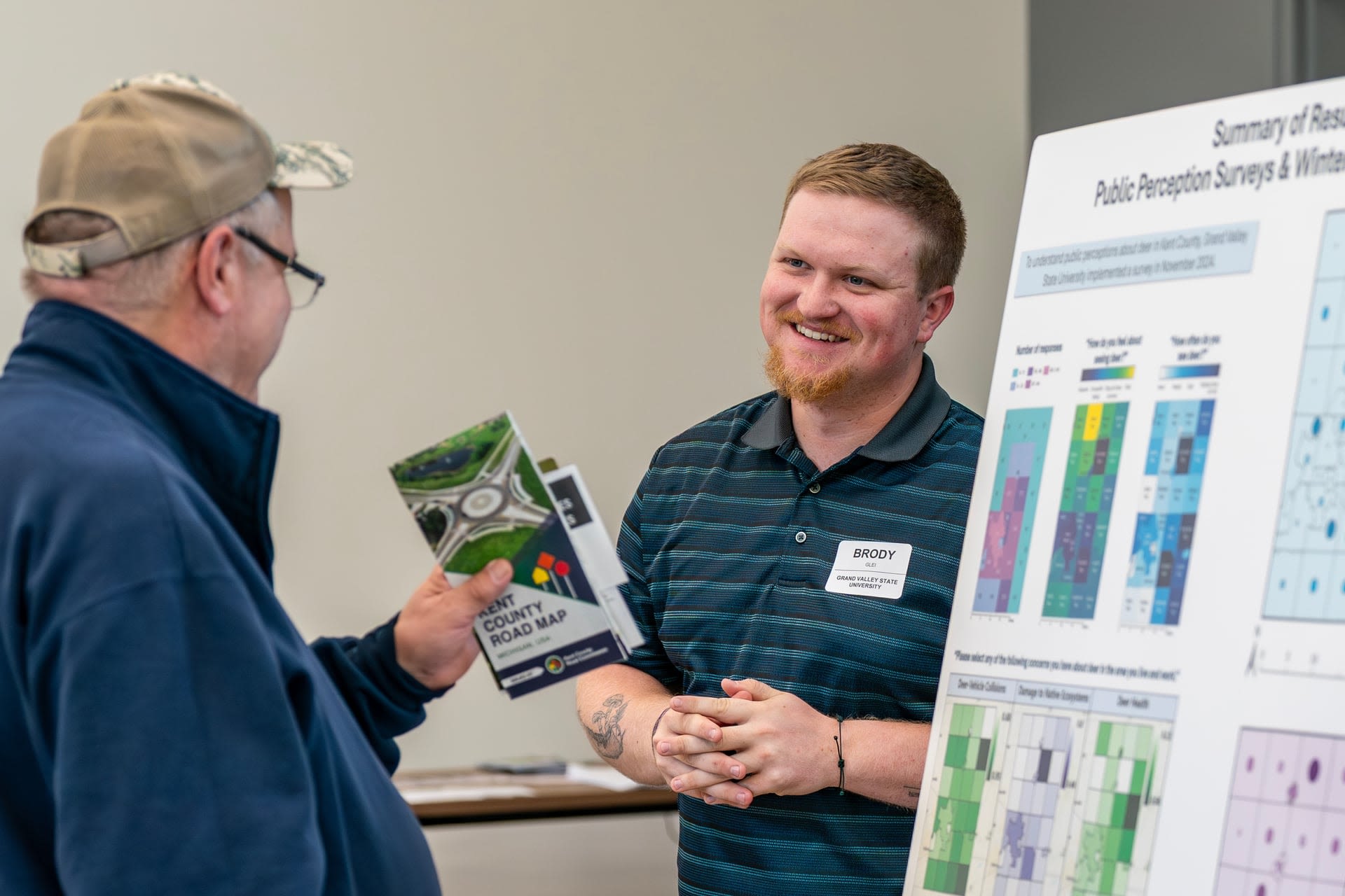 Brody Glei smiles while standing by a colorful presentation board as he talks to a Eric Vander Stel holding a pamphlet up in his hand.