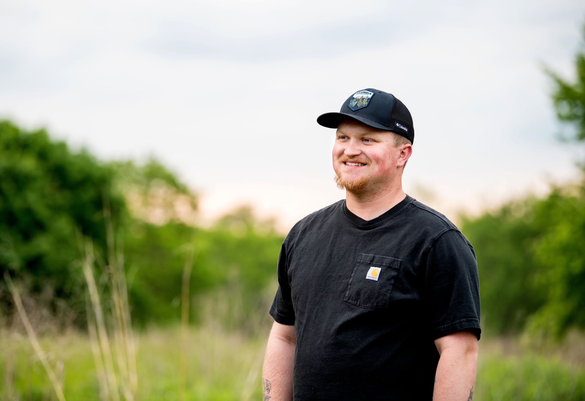 Brody Glei smiles while standing outside in front of a lush green landscape.