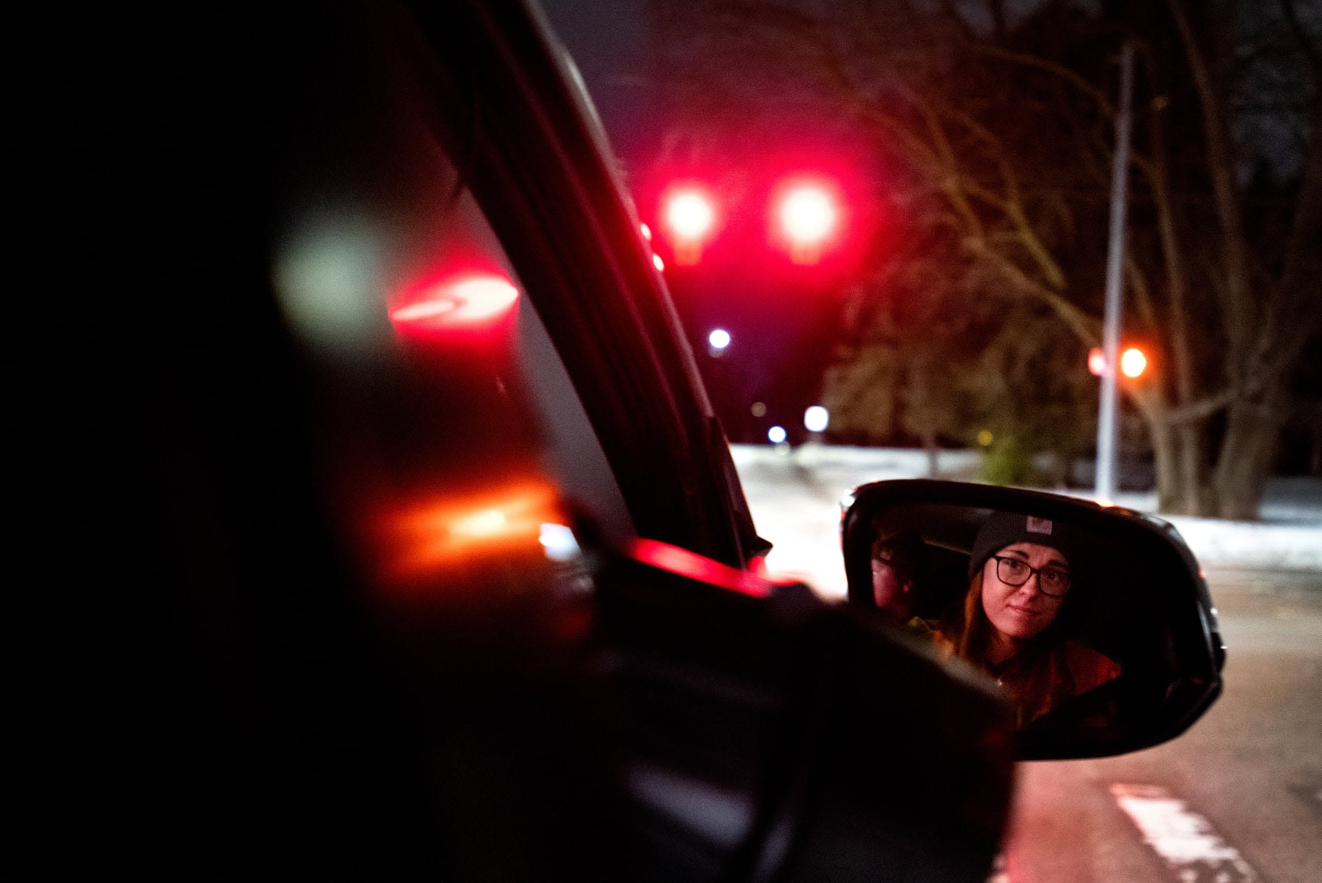 A close-up view from the inside of a car, showing a woman with a beanie looking in the side mirror, with red traffic light illuminating the background.