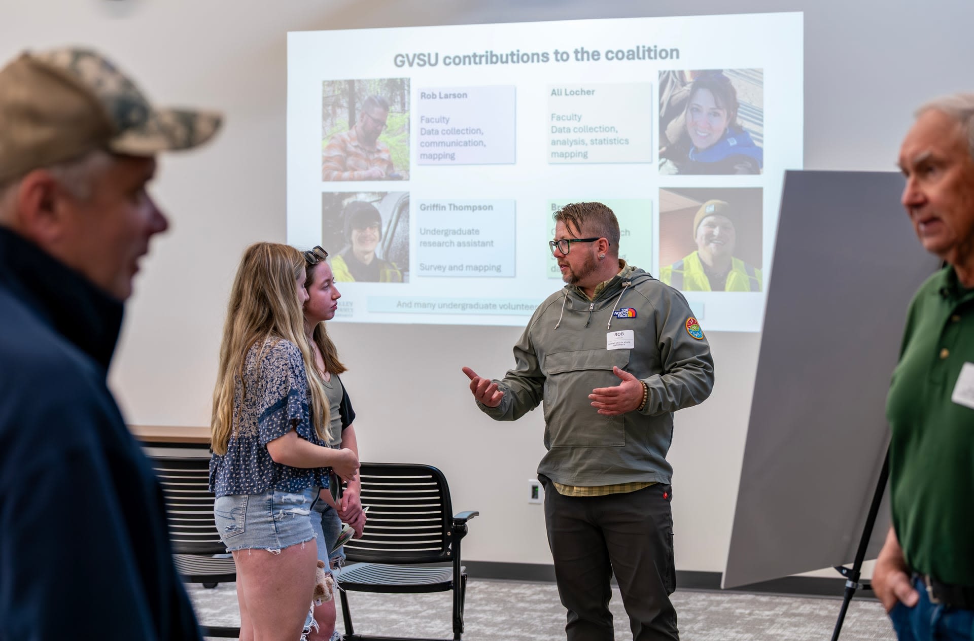 Three individuals stand conversing in front of a presentation on deer-related vehicle collision risks are projected on the wall. 